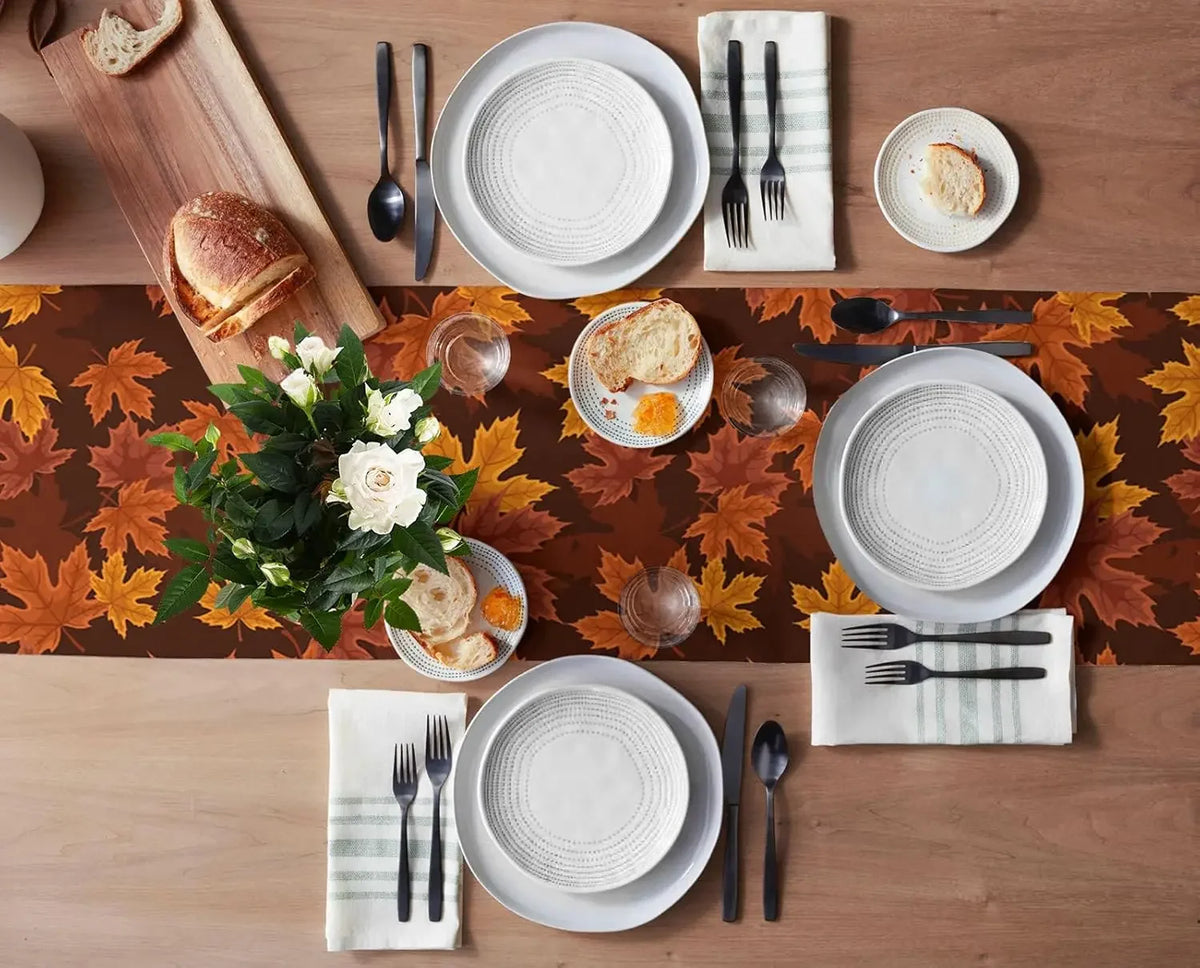 Cette image montre une table de petit-déjeuner automnale avec des assiettes, des ustensiles et des accessoires de table sur un fond de feuilles d'érable orangées, accompagnée d'un bouquet de fleurs blanches.