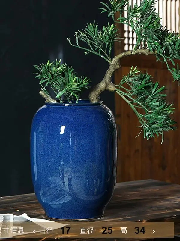 Vase en céramique bleu foncé avec des rameaux de plantes vertes, sur une surface en bois sombre.