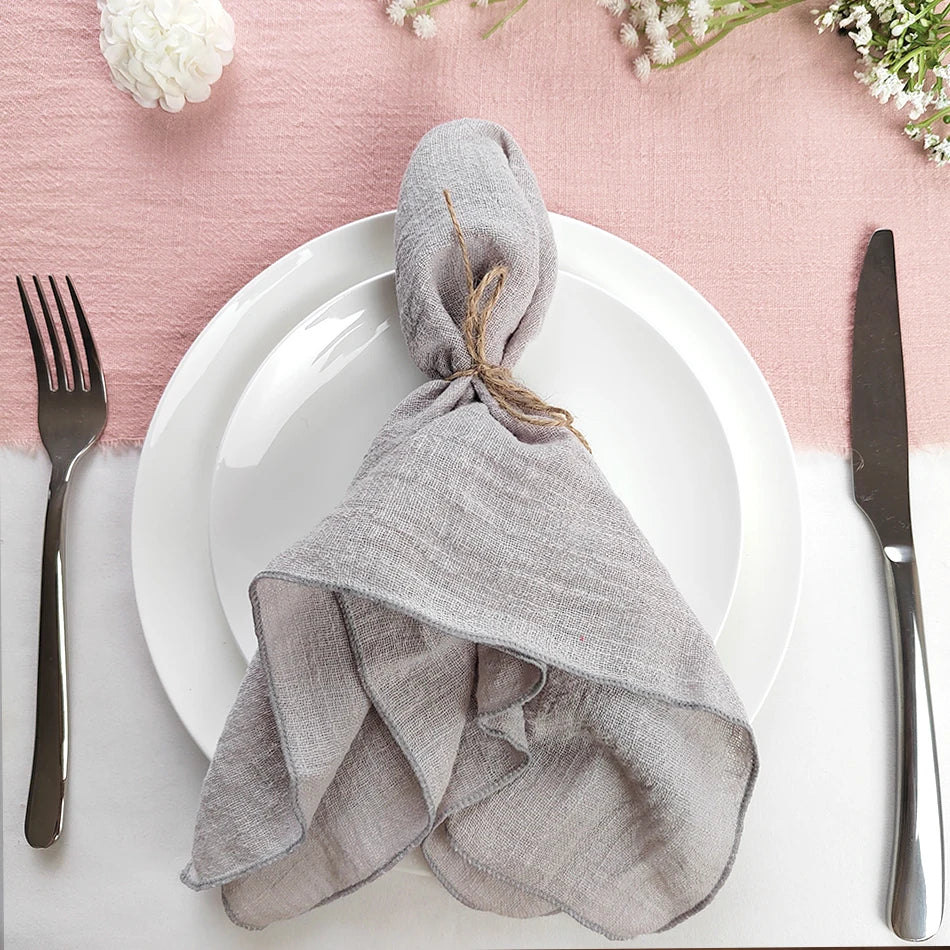 Assiette blanche avec serviette grise élégamment nouée, couteau et fourchette noirs, sur fond rose pastel orné de fleurs blanches.