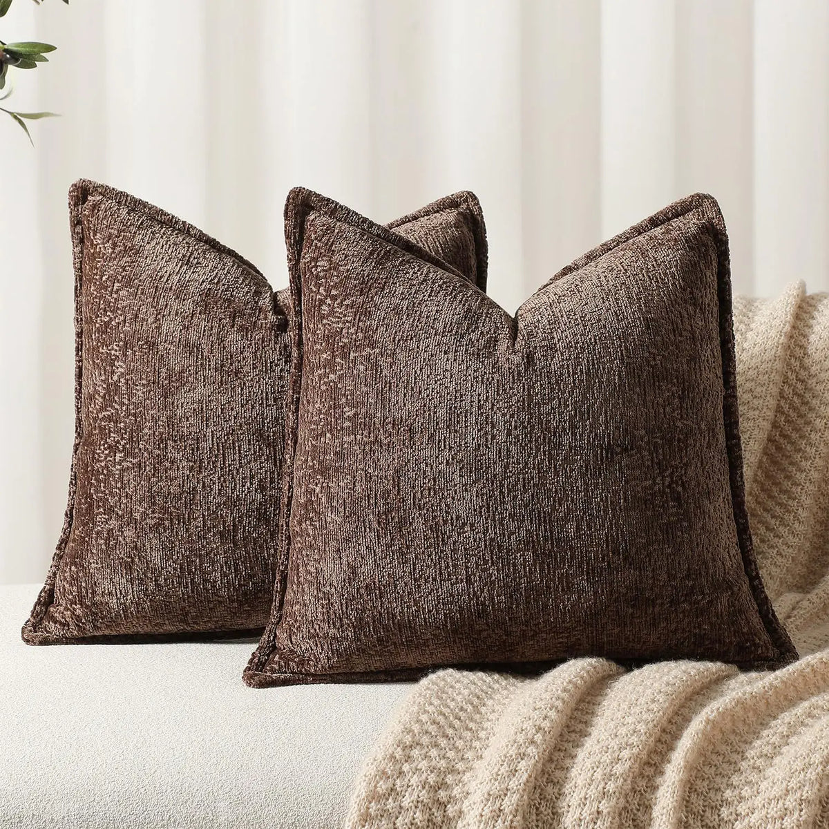 Trois coussins confortables en velours brun foncé, avec des motifs texturés, reposent sur une couverture beige moelleuse, créant une atmosphère chaleureuse et accueillante dans l'espace.