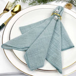 Serviette grise délicatement nouée avec des détails dorés, posée élégamment sur une assiette blanche avec des couverts dorés, le tout accompagné d'éléments végétaux.