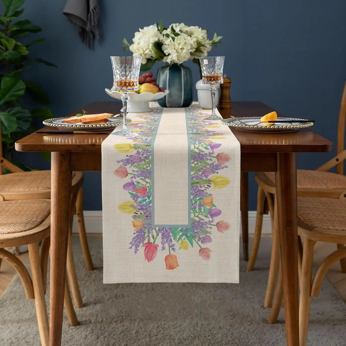 Une table de salle à manger élégante avec un chemin de table fleuri, des verres à vin et des fleurs dans un vase.