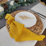 Une assiette blanche posée sur un napperon tressé, entourée de décorations végétales automnales, avec un beau linge de table jaune.