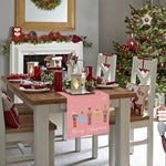 Cette image présente une table de salle à manger joliment décorée pour Noël, avec des ornements rouges et verts, des bougies, et un arbre de Noël dans un coin. La table est recouverte d'un chemin de table avec des motifs de personnages de Noël et l'inscription "Joyeux Noël".
