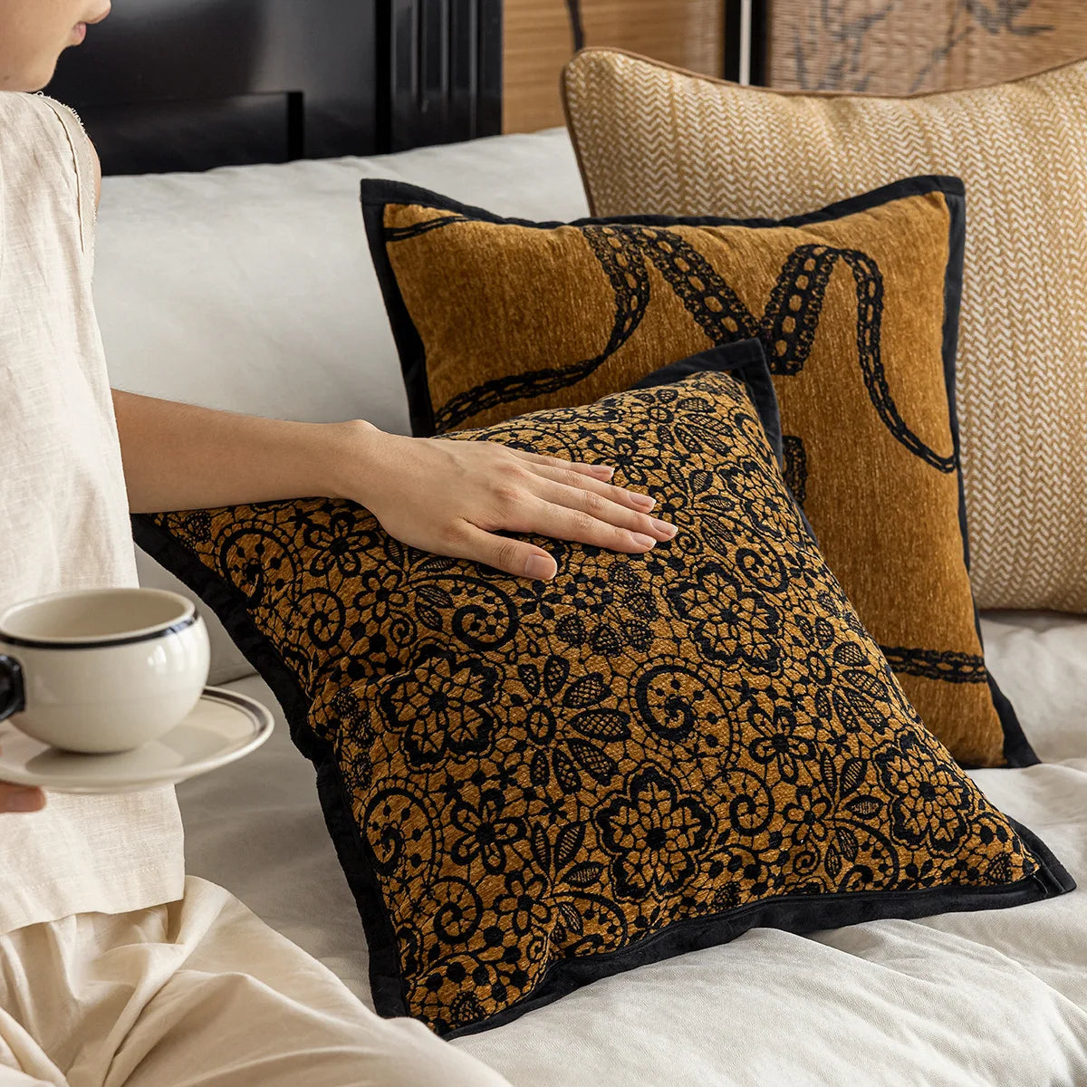Coussins de canapé en tissu jaune moutarde avec motifs floraux noirs, disposés sur un lit blanc avec une tasse de café sur la table de chevet.