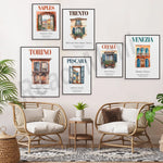 Une série d'affiches vintage présentant différentes villes italiennes comme Naples, Trento, Torino, Pescara et Venise, accrochées au mur d'un salon chaleureux et confortable meublé de fauteuils en rotin.
