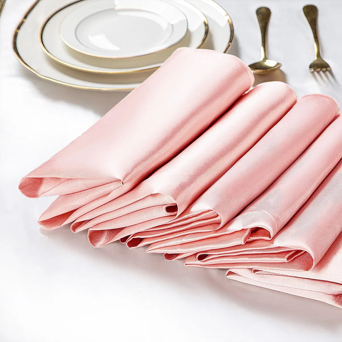 Serviettes en tissu rose élégantes et pliées avec des assiettes et des couverts dorés sur fond clair.
