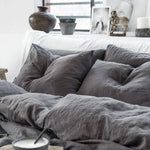 Cette image présente un lit douillet recouvert de draps et de coussins gris, créant une atmosphère apaisante et cocooning dans la pièce, avec des éléments décoratifs comme des vases et des statues disposés sur une étagère.