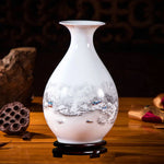 Un vase en porcelaine blanche orné d'une scène hivernale peinte, avec des maisons dans un paysage enneigé, sur un support en bois.
