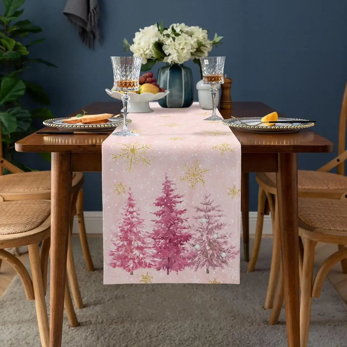 Table décorée avec un centre de table composé d'un vase de fleurs blanches, de verres à vin et de fruits, sur un chemin de table imprimé de motifs arborés hivernaux dans des tons roses et dorés.
