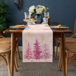 Table décorée avec un centre de table composé d'un vase de fleurs blanches, de verres à vin et de fruits, sur un chemin de table imprimé de motifs arborés hivernaux dans des tons roses et dorés.