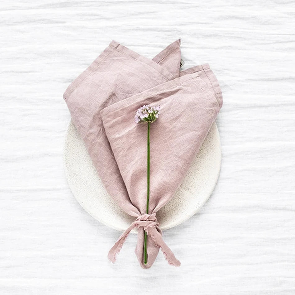 Une serviette de table en lin rose pâle avec une petite fleur blanche au centre, posée sur un fond blanc.