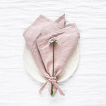 Une serviette de table en lin rose pâle avec une petite fleur blanche au centre, posée sur un fond blanc.