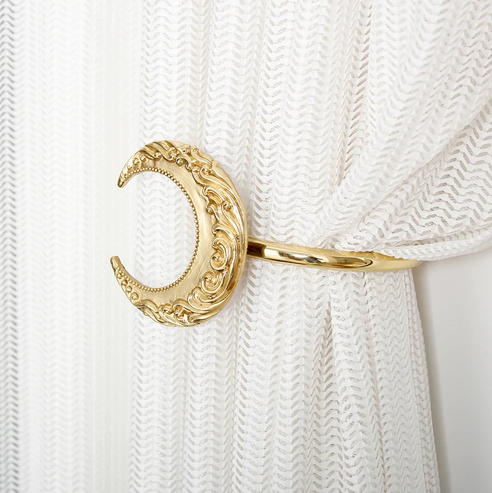 Un élégant élément de rideau décoratif en forme de lune dorée, ornée de motifs sculptés et suspendu sur un rideau blanc.