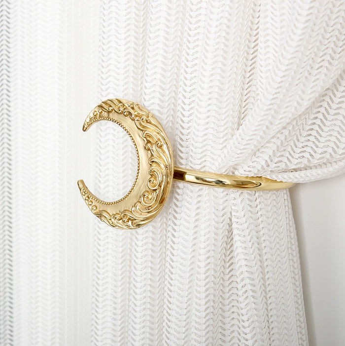 Un élégant élément de rideau décoratif en forme de lune dorée, ornée de motifs sculptés et suspendu sur un rideau blanc.
