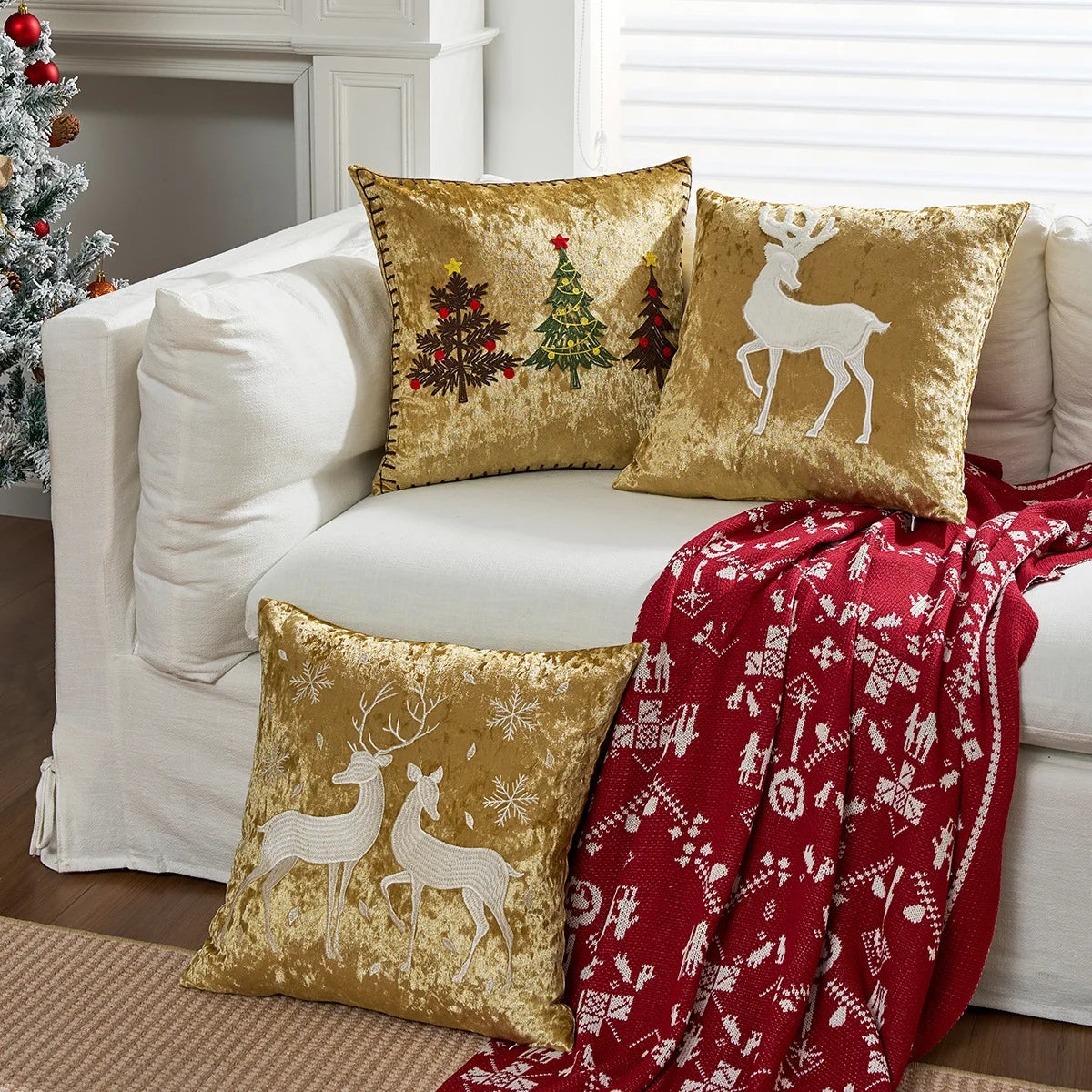 Des coussins décoratifs dorés avec des motifs de sapins de Noël et de cerfs, posés sur un canapé blanc, accompagnés d'un plaid en tricot rouge avec des motifs de flocons de neige, créant une ambiance festive et chaleureuse dans la pièce.