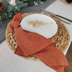Une assiette blanche avec un motif doré posée sur un napperon tressé en fibres naturelles, accompagnée d'un linge de table en coton rouille.