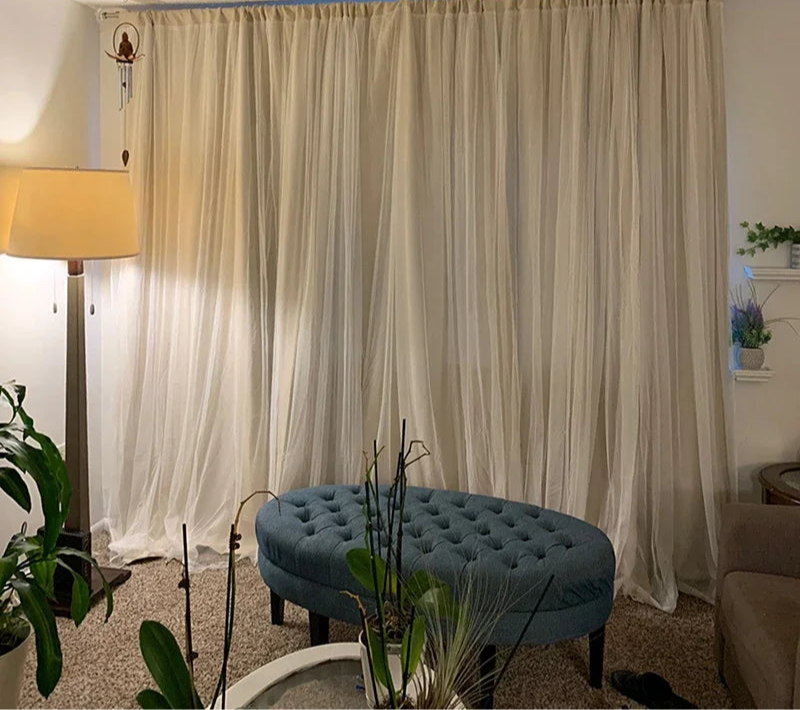 Salon moderne avec rideaux translucides beige créant une ambiance douce. Grand pouf bleu capitonné en avant-plan, entouré de plantes vertes. Lampe d'appoint à gauche offrant un éclairage chaleureux. Idéal pour un espace de détente ou de lecture.