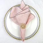 Serviette de table pliée en éventail rose pâle avec épingle décorative en métal doré en forme de fleur posée sur un plateau de service rond orné de perles dorées.