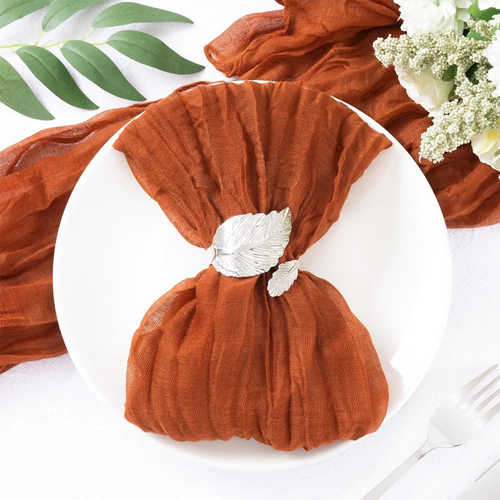 Un foulard en tissu plissé de couleur rouille, décoré d'un élégant fermoir en feuille métallique, posé sur un fond blanc avec des feuilles vertes et des fleurs blanches.