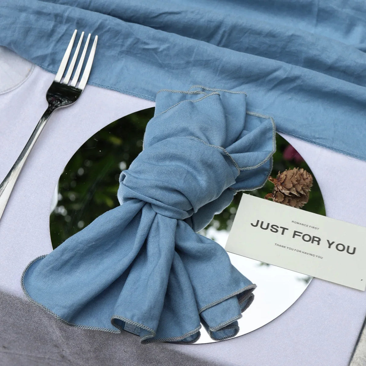 Une serviette en tissu bleu clair est enroulée de manière artistique sur un plat blanc, avec une fourchette métallique à côté et une petite carte avec le texte "Just for you" posée sur le dessus.