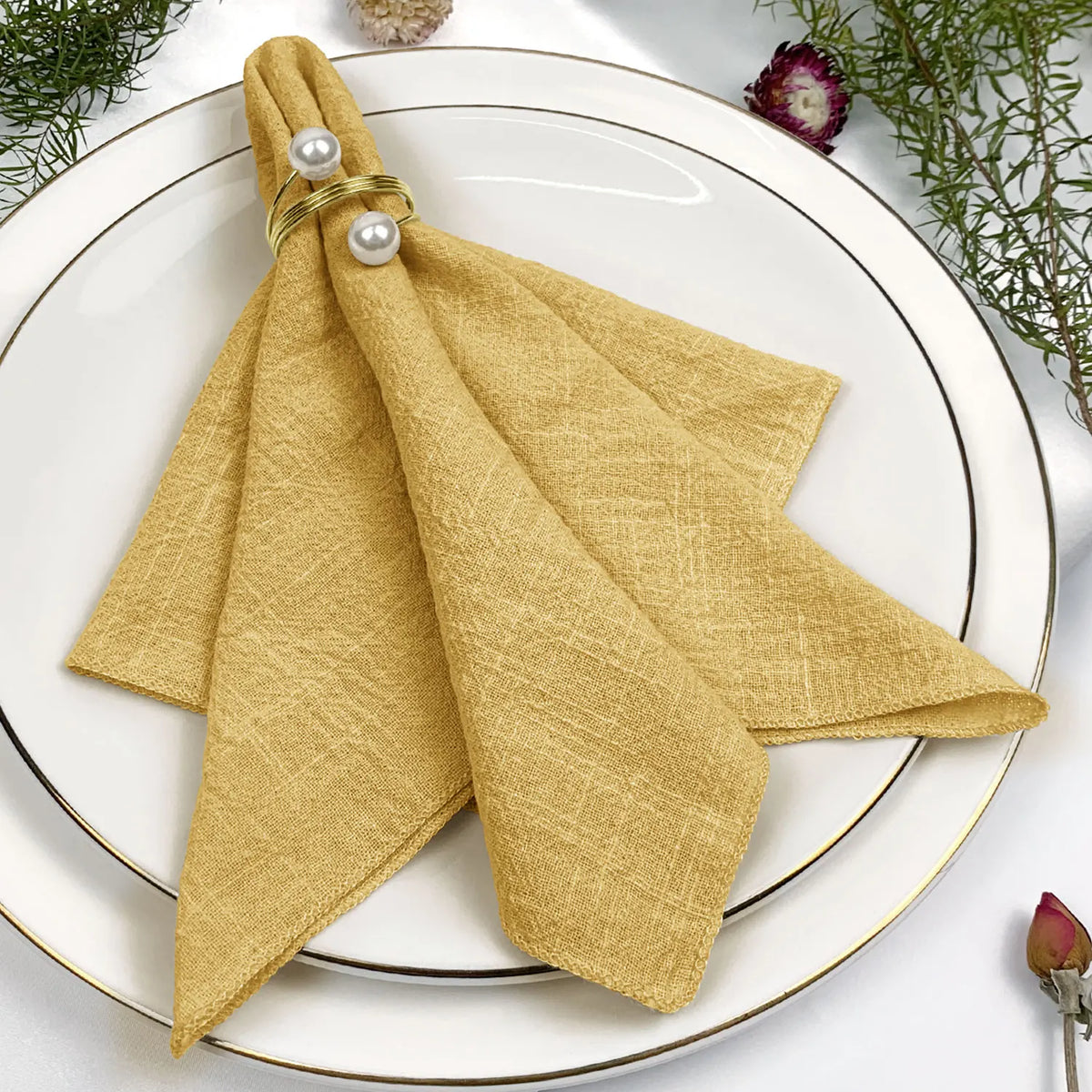 Serviette de table pliée de couleur dorée, ornée de perles, posée sur une assiette blanche entourée de fleurs.