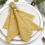 Serviette de table pliée de couleur dorée, ornée de perles, posée sur une assiette blanche entourée de fleurs.