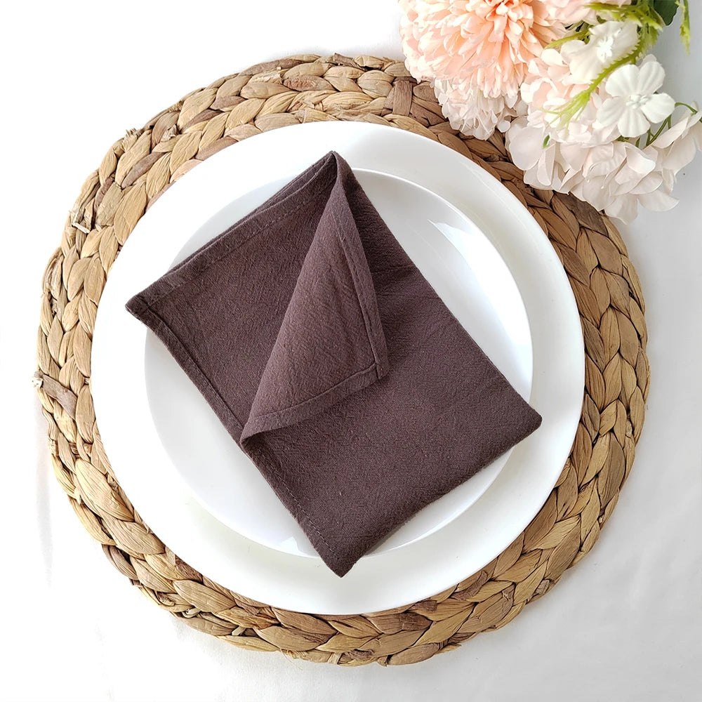 Une serviette de table en tissu brun foncé repose sur une assiette blanche au centre d'un dessous de plat en fibres tressées, entouré de fleurs roses et blanches.