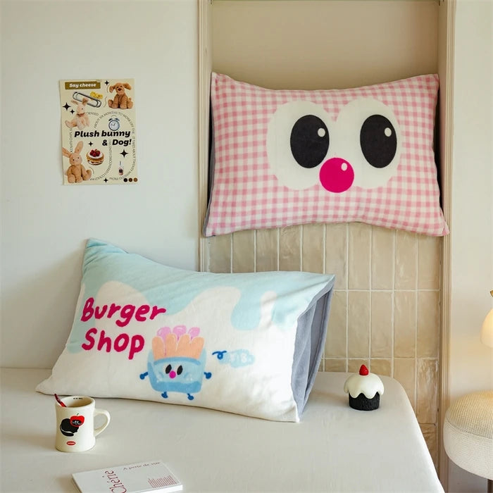 Coussin en forme d'yeux avec un nez rose sur un lit, ainsi qu'un oreiller avec un personnage de dessin animé représentant une boutique de hamburgers.