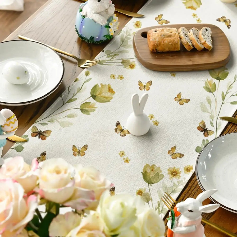 Sur la table, on trouve des décorations de Pâques fleuries avec des papillons dorés, des plats en porcelaine, des ustensiles en bois et des pâtisseries maison.