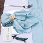 Une serviette de table en tissu bleu pâle décorée d'une broche ornée de perles et de cristaux, accompagnée d'une fourchette métallique posée à côté.
