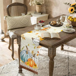 Une table de cuisine rustique avec des décorations d'automne, une serviette imprimée avec des citrouilles et des fleurs, un livre ouvert et un arrangement floral dans un vase.