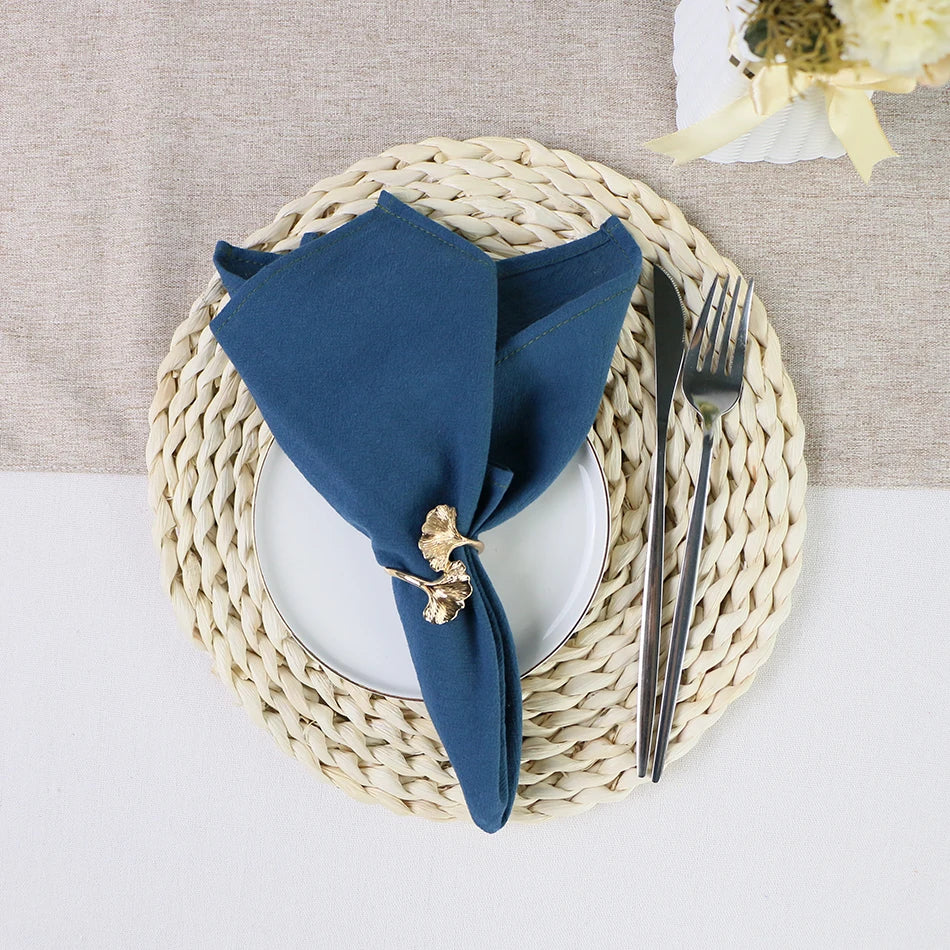 Un élégant ensemble de table composé d'un napperon tressé beige, d'une assiette blanche et d'une serviette de table bleu marine décorée d'un élément doré.