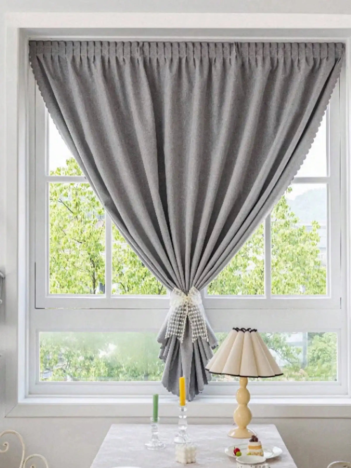 Rideau gris élégant ornant une fenêtre de salon, laissant passer la lumière naturelle. Décoration intérieure moderne avec lampe et bougies sur une table, créant une ambiance chaleureuse et accueillante. Vue sur jardin en arrière-plan.