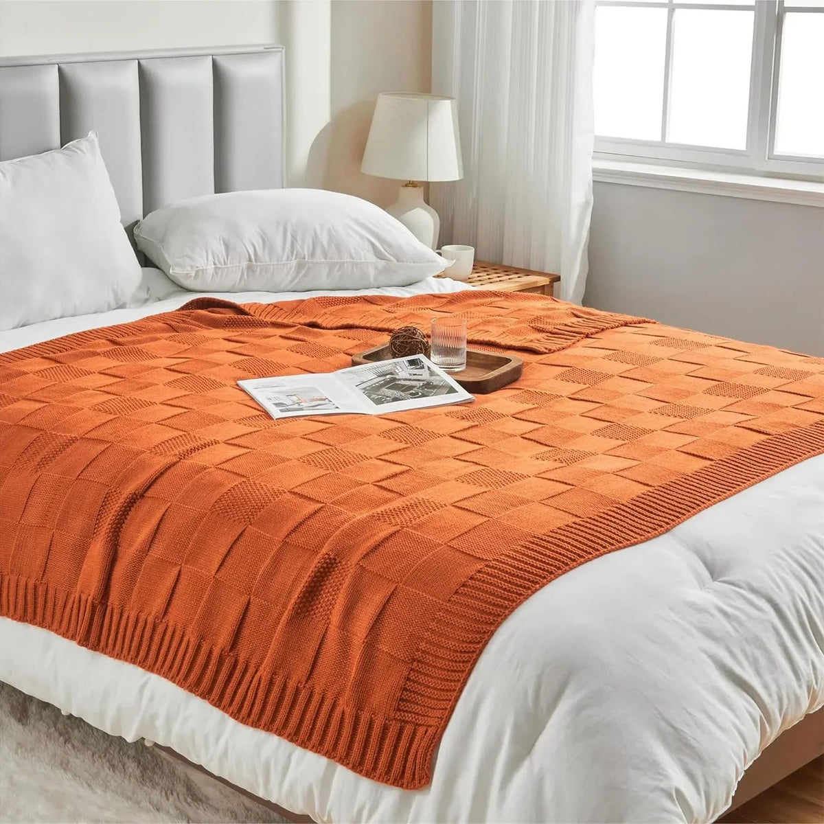 Couverture en maille orange carrée posée sur un lit blanc dans une chambre avec rideaux et lampe de chevet.