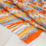 Une couverture tissée multicolore aux motifs géométriques vibrants et aux franges frangées, dans des tons chauds d'orange, de bleu et de jaune.