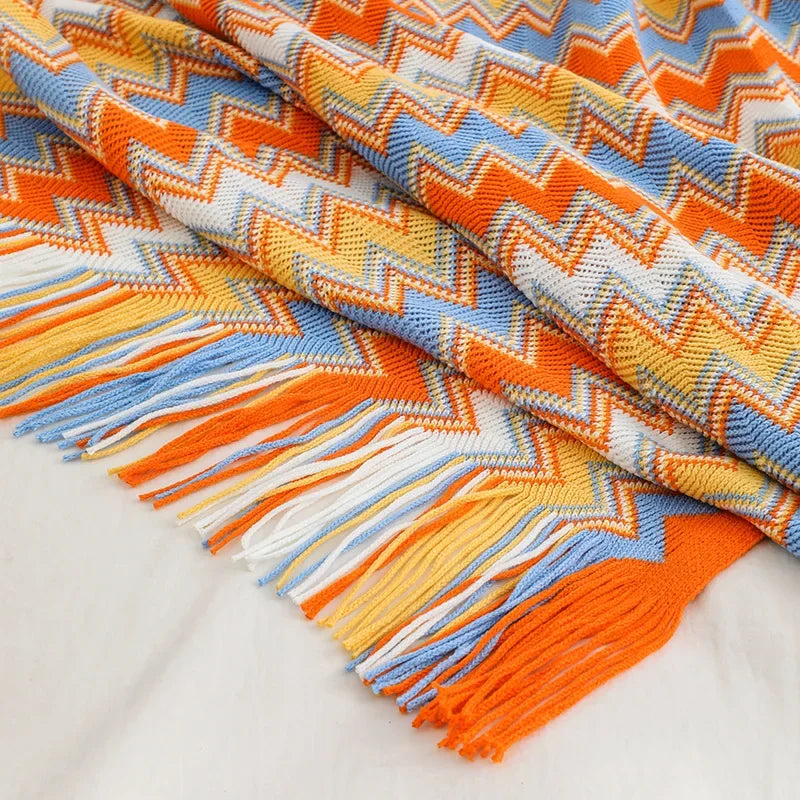 Une couverture tissée multicolore aux motifs géométriques vibrants et aux franges frangées, dans des tons chauds d'orange, de bleu et de jaune.