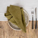 Des serviettes de table en lin vert et une assiette en pierre posées sur une table en bois rustique, avec des couverts en métal.