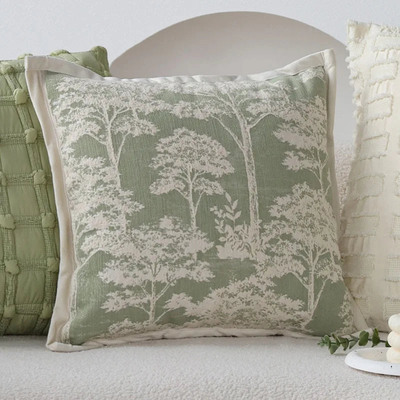 L'image montre un oreiller décoratif de forme carrée, avec un motif de silhouettes d'arbres dans des tons verts et blancs, créant une atmosphère apaisante et naturelle dans la pièce.