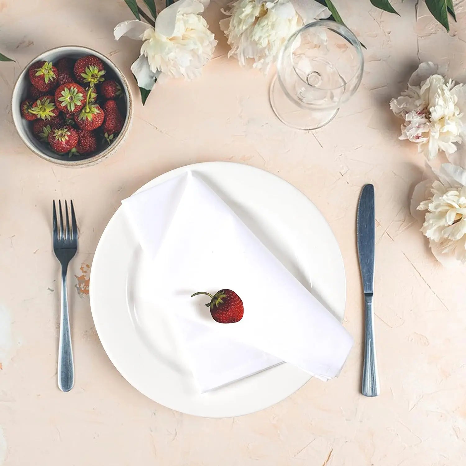 L'image montre une belle table de fête avec une assiette blanche, une fourchette et un couteau posés à côté d'un bol de fraises fraîches. Des fleurs blanches ornent la scène, créant une atmosphère agréable et élégante.