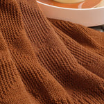 Un plaid en tricot de laine d'un riche ton de rouille, avec un motif de points et de rayures, donnant une impression douce et confortable.