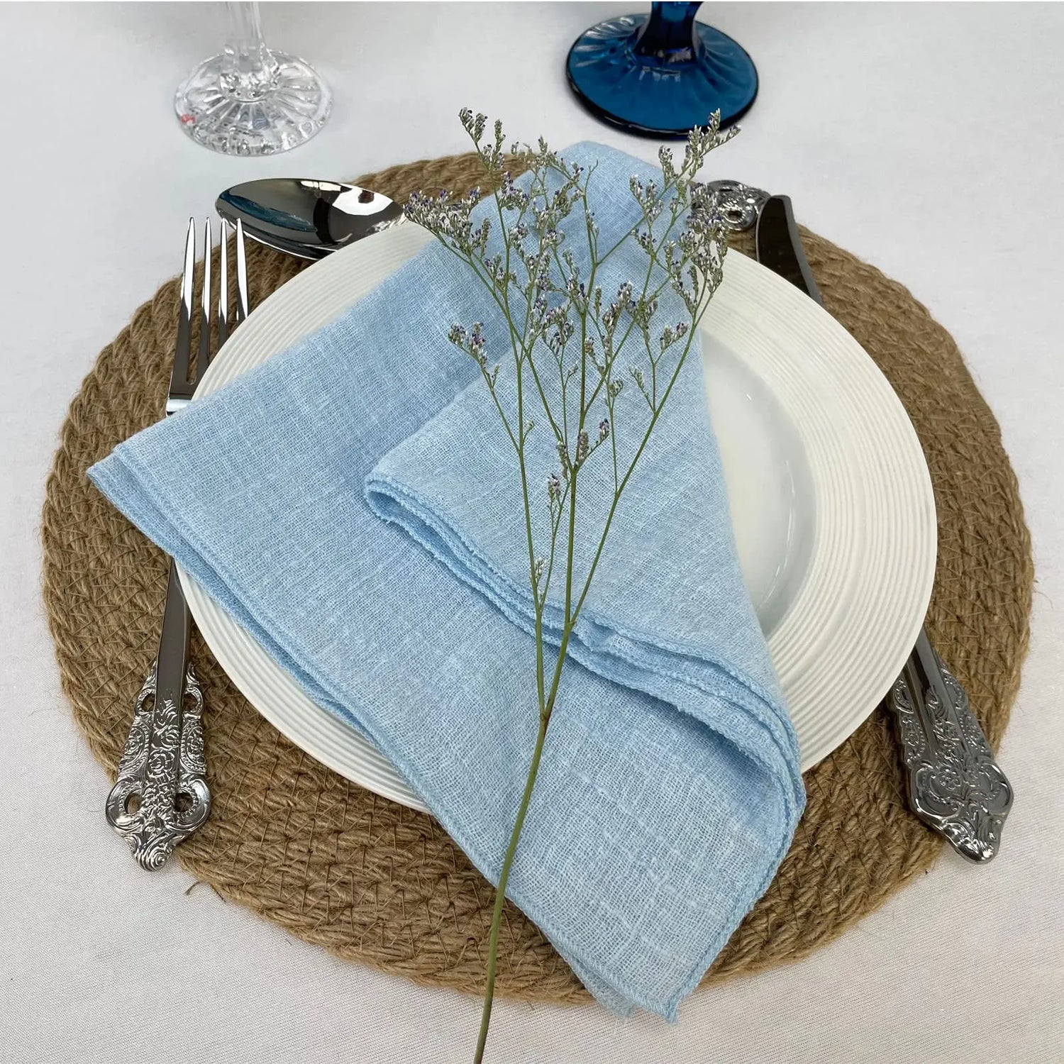 Une table élégamment dressée avec des accessoires en verre, des couverts en argent et une serviette en lin bleu ciel accentuée par une touche florale délicate.
