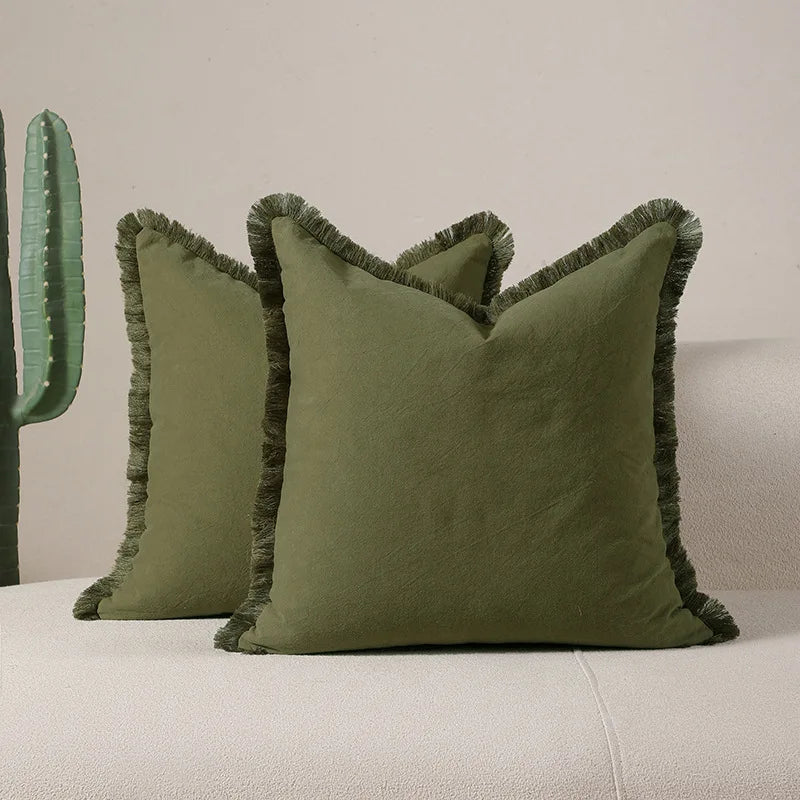 Deux oreillers carrés vert olive avec une bordure de franges vertes sont posés sur un canapé blanc.