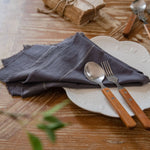 L'image montre un arrangement de table avec des couverts en acier inoxydable et un linge de table gris sur un fond en bois, suggérant un cadre rustique et naturel pour un repas.