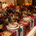 Cette image montre une belle table de Noël décorée avec une composition florale de brindilles de sapin, de baies rouges et de bougies allumées, ainsi que des cadeaux emballés dans du papier cadeau rouge et blanc festif.