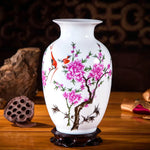 Un vase en porcelaine blanche décoré de fleurs de cerisier roses épanouies et d'oiseaux rouges sur un socle en bois sombre.