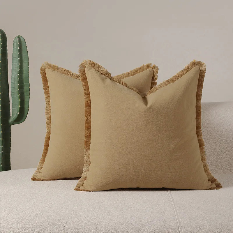 Deux oreillers décoratifs de couleur beige clair avec des bordures en franges, posés sur un canapé blanc à côté de plantes vertes.