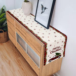 Sur un meuble en bois clair avec des tiroirs, un chemin de table aux motifs de flocons de neige et de voitures rouges vintage transmet une ambiance festive de Noël.