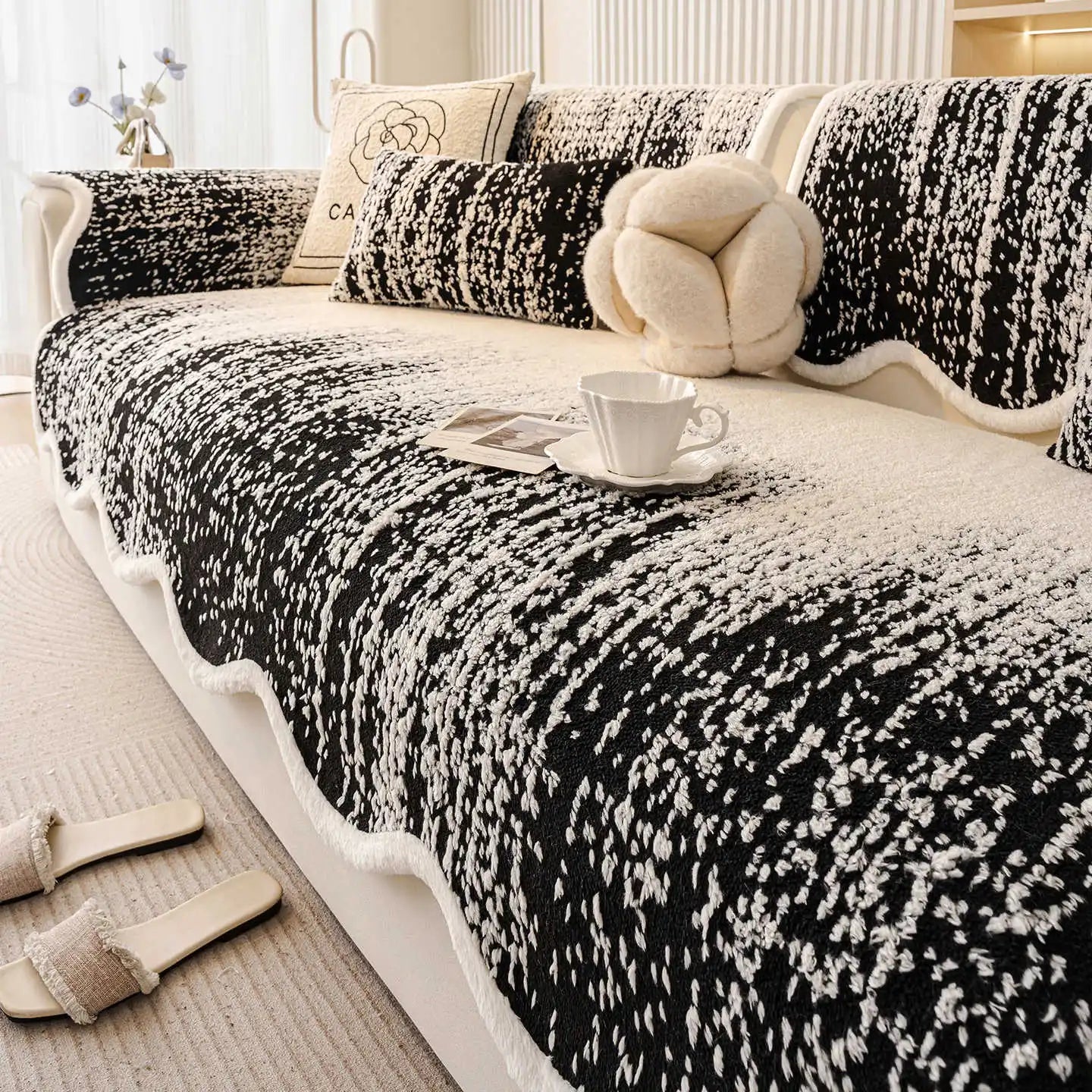 Couverture de canapé noire et blanche avec des motifs abstraits et un ours en peluche posé sur le côté, avec une tasse de café sur une table basse.