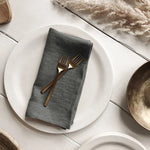 Assiette blanche avec serviette grise et couverts en métal doré sur une table en bois clair, ornée de décorations en feuillage sec.